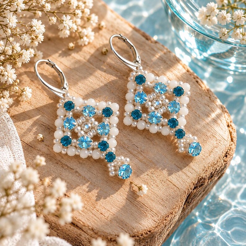 May include: A pair of dangle earrings featuring a square geometric pattern crafted from small white beads and alternating light and dark blue crystals. Each earring is finished with a silver-toned leverback closure and a single blue crystal drop.