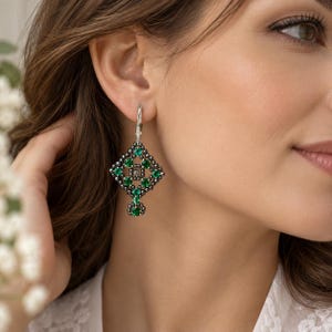 May include: A drop earring featuring a diamond-shaped lattice design crafted from small, dark metallic beads. The geometric structure is accented with multiple round, vibrant green gemstones and a single clear stone at the center.