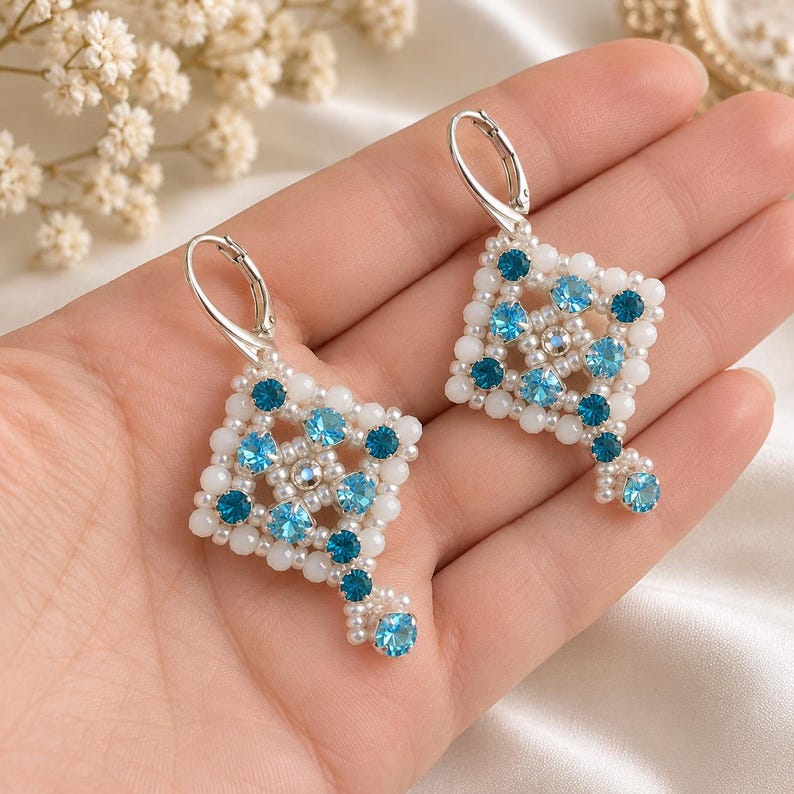 May include: A pair of dangle earrings featuring a diamond-shaped geometric pattern crafted from small white beads and faceted light blue and dark teal crystals. The earrings are finished with silver-toned lever-back closures for secure wear.