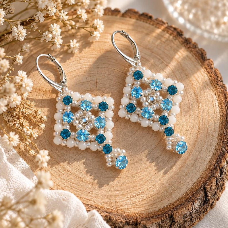 May include: A pair of dangle earrings featuring a square geometric pattern crafted from small white beads and round blue crystals. The earrings are finished with silver-toned leverback closures, creating a structured design with alternating light and dark blue accents.