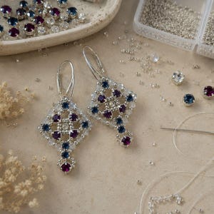 May include: A pair of dangle earrings featuring a diamond-shaped lattice design crafted from small silver beads, deep purple round crystals, and dark blue round crystals. Each earring is suspended from a silver-toned metal leverback closure.