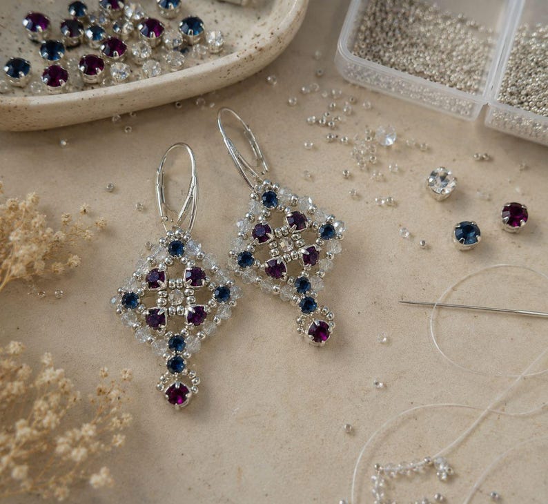 May include: A pair of dangle earrings featuring a diamond-shaped lattice design crafted from small silver beads, deep purple round crystals, and dark blue round crystals. Each earring is suspended from a silver-toned metal leverback closure.