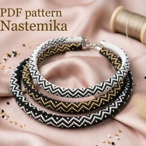 May include: Three beaded necklaces with a chevron pattern in black, white, and gold. The necklaces are displayed on a pink satin surface. The text "PDF pattern Nastemika" is in the upper left corner.