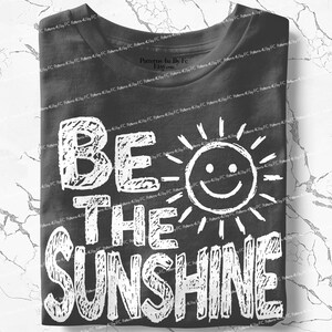 May include: A folded charcoal gray t-shirt with the words "BE THE SUNSHINE" in large, white, distressed lettering. A smiling sun graphic is incorporated into the design. The shirt is made of soft material.
