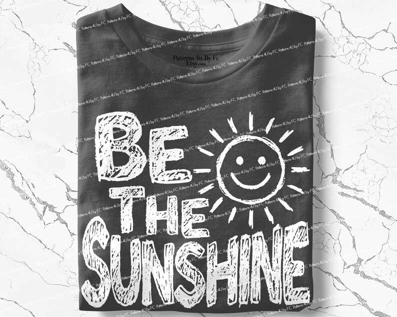 May include: A folded charcoal gray t-shirt with the words "BE THE SUNSHINE" in large, white, distressed lettering. A smiling sun graphic is incorporated into the design. The shirt is made of soft material.