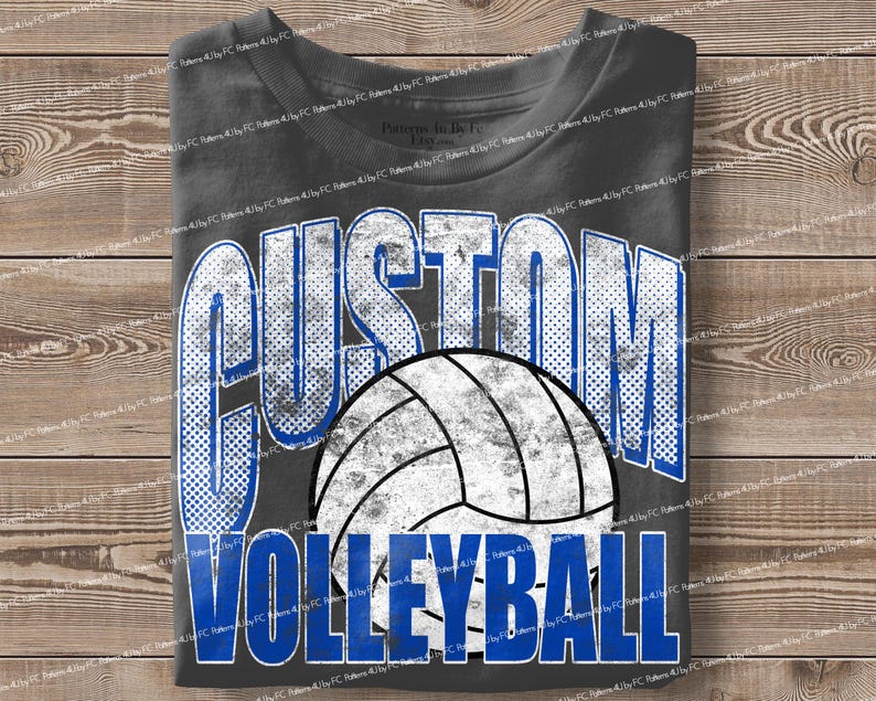 May include: A charcoal gray t-shirt featuring the words "CUSTOM VOLLEYBALL" in a distressed, blue and white design. A volleyball graphic is centered below the word "CUSTOM". The shirt is folded on a wooden surface.
