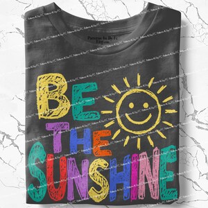 May include: A folded charcoal gray t-shirt with the words "BE THE SUNSHINE" in colorful, hand-drawn letters. A smiling sun graphic is incorporated into the design, adding a cheerful touch to the apparel.