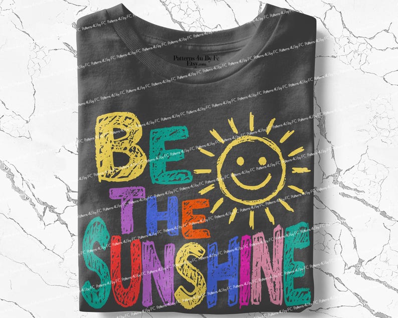 May include: A folded charcoal gray t-shirt with the words "BE THE SUNSHINE" in colorful, hand-drawn letters. A smiling sun graphic is incorporated into the design, adding a cheerful touch to the apparel.
