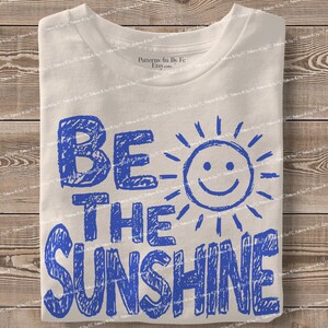 May include: A folded, off-white t-shirt with the words "BE THE SUNSHINE" in blue, handwritten-style lettering. A smiling sun graphic is next to the word "BE". The shirt is on a wooden surface.