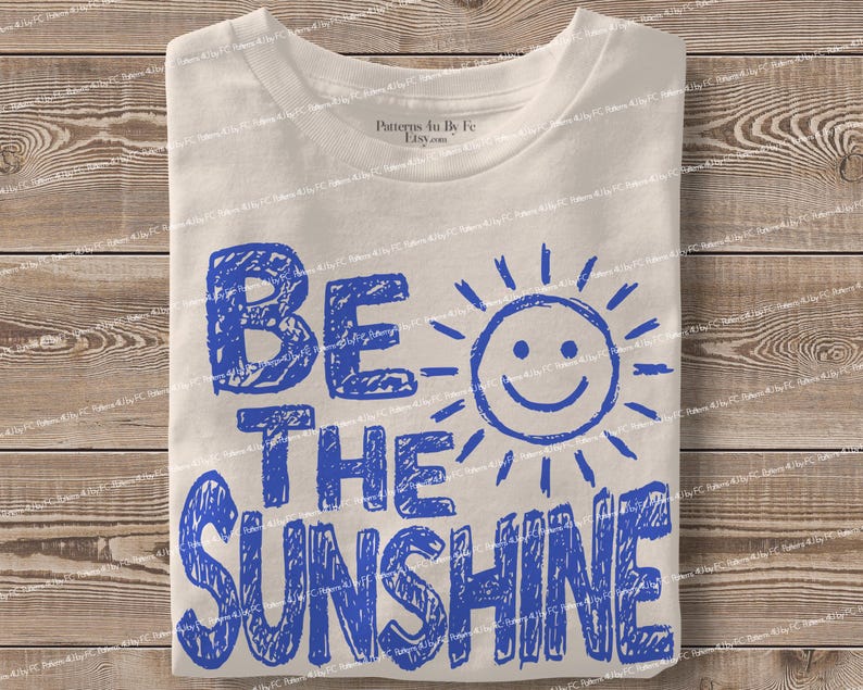May include: A folded, off-white t-shirt with the words "BE THE SUNSHINE" in blue, handwritten-style lettering. A smiling sun graphic is next to the word "BE". The shirt is on a wooden surface.