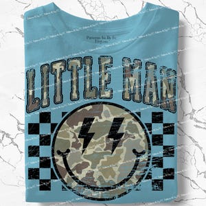 K&ouml;nnte beinhalten: Hellblaues T-Shirt mit dem Text "LITTLE MAN" in einem Camouflage-Design. Darunter befindet sich ein Smiley mit Camouflage-Muster und Blitzaugen, vor einem schwarz-wei&szlig;en Schachbrettmuster.