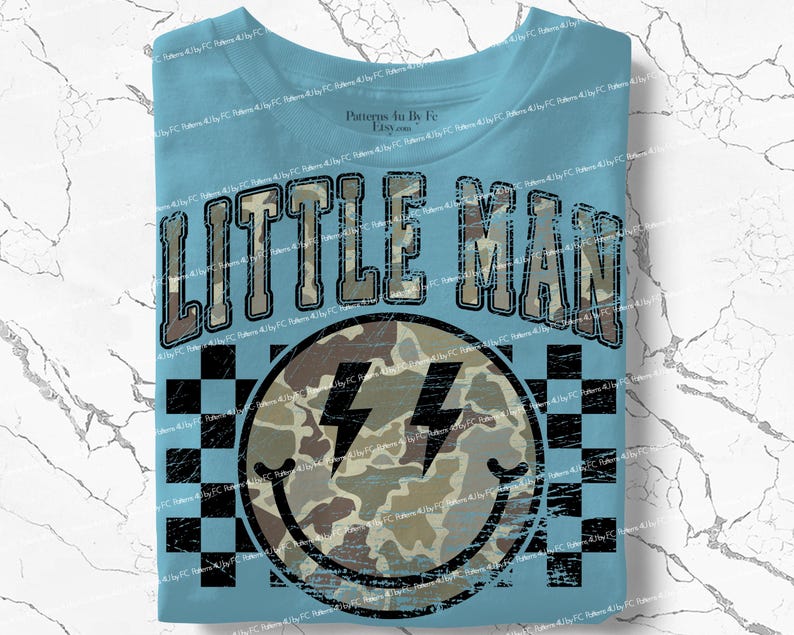 K&ouml;nnte beinhalten: Hellblaues T-Shirt mit dem Text "LITTLE MAN" in einem Camouflage-Design. Darunter befindet sich ein Smiley mit Camouflage-Muster und Blitzaugen, vor einem schwarz-wei&szlig;en Schachbrettmuster.