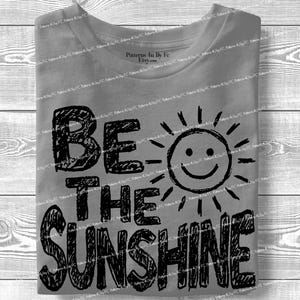 May include: Gray t-shirt with the phrase "BE THE SUNSHINE" in black, handwritten-style lettering. A smiling sun graphic is next to the word "SUNSHINE". The shirt is folded neatly.