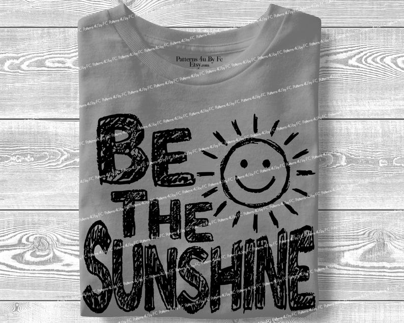 May include: Gray t-shirt with the phrase "BE THE SUNSHINE" in black, handwritten-style lettering. A smiling sun graphic is next to the word "SUNSHINE". The shirt is folded neatly.