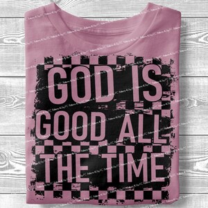May include: A dusty rose t-shirt with the text "GOD IS GOOD ALL THE TIME" in large black letters. The text is set against a black and white checkered background, creating a bold contrast. The shirt is folded neatly.