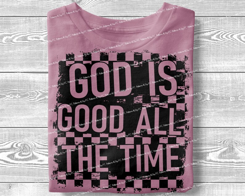 May include: A dusty rose t-shirt with the text "GOD IS GOOD ALL THE TIME" in large black letters. The text is set against a black and white checkered background, creating a bold contrast. The shirt is folded neatly.