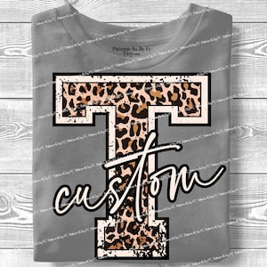 Op de afbeelding: Grijs T-shirt met een grote, op maat gemaakte letter "T" met een luipaardprint. Het woord "custom" staat eronder in een sierlijk lettertype. Het shirt wordt gepresenteerd op een houten oppervlak.
