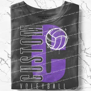 May include: A black t-shirt with a large purple "G" and a volleyball graphic. The word "CUSTOM" is printed vertically on the left side, and "VOLLEYBALL" is printed at the bottom in white.