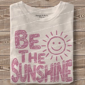 May include: A folded white t-shirt with the phrase "BE THE SUNSHINE" in pink block letters and a smiling sun graphic. The shirt is made of a soft material and is folded neatly.