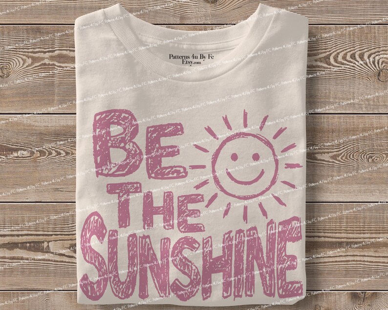 May include: A folded white t-shirt with the phrase "BE THE SUNSHINE" in pink block letters and a smiling sun graphic. The shirt is made of a soft material and is folded neatly.