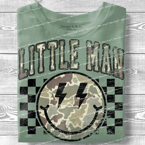 K&ouml;nnte beinhalten: Salbeigr&uuml;nes T-Shirt mit dem Text "LITTLE MAN" in einem Camouflage-Design. Darunter befindet sich ein Smiley mit Camouflage-Muster und zwei schwarzen Blitzen. Der Hintergrund hat ein schwarz-wei&szlig;es Schachbrettmuster.