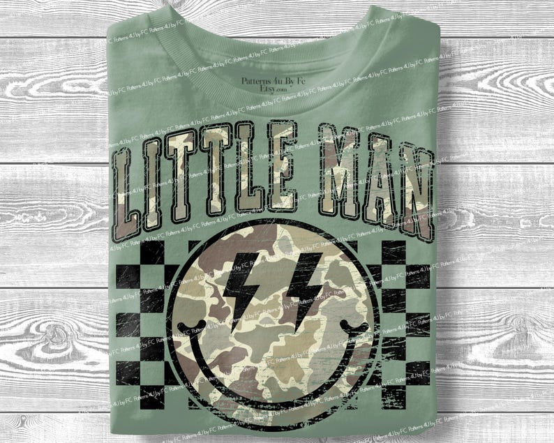 K&ouml;nnte beinhalten: Salbeigr&uuml;nes T-Shirt mit dem Text "LITTLE MAN" in einem Camouflage-Design. Darunter befindet sich ein Smiley mit Camouflage-Muster und zwei schwarzen Blitzen. Der Hintergrund hat ein schwarz-wei&szlig;es Schachbrettmuster.