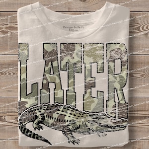 May include: Cream-coloured t-shirt featuring the word "LATER" in large, distressed, camouflage-patterned letters. An alligator illustration is below the text. The shirt is displayed on a wooden surface.