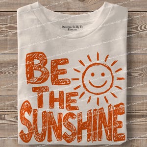 May include: A folded, off-white t-shirt with the words "BE THE SUNSHINE" in orange block letters and a smiling sun graphic. The shirt is on a wooden surface.