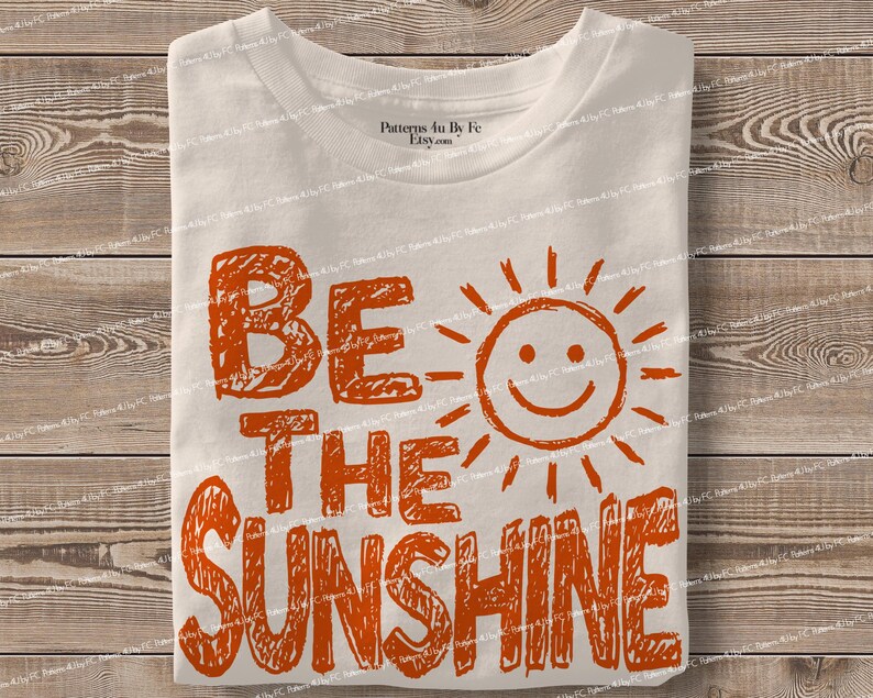 May include: A folded, off-white t-shirt with the words "BE THE SUNSHINE" in orange block letters and a smiling sun graphic. The shirt is on a wooden surface.