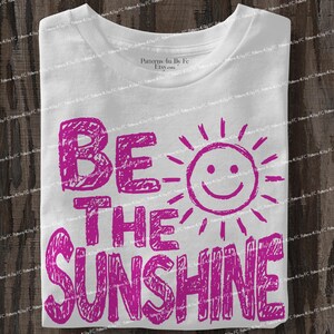 May include: White t-shirt with the phrase "BE THE SUNSHINE" in bright pink, accompanied by a smiling sun graphic. The text and graphic have a hand-drawn, textured appearance, creating a cheerful and playful design.