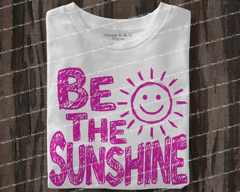 May include: White t-shirt with the phrase "BE THE SUNSHINE" in bright pink, accompanied by a smiling sun graphic. The text and graphic have a hand-drawn, textured appearance, creating a cheerful and playful design.