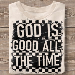 May include: Cream-colored t-shirt with the text "GOD IS GOOD ALL THE TIME" in bold, black letters. The text is set against a black and white checkered background. The shirt is folded on a wooden surface.