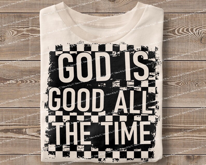 May include: Cream-colored t-shirt with the text "GOD IS GOOD ALL THE TIME" in bold, black letters. The text is set against a black and white checkered background. The shirt is folded on a wooden surface.