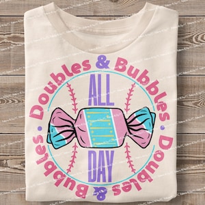 Puede incluir: Una camiseta blanca con un diseño gráfico rosa, azul y amarillo. El gráfico presenta una pelota de béisbol con el texto "Doubles & Bubbles All Day Doubles & Bubbles" alrededor. Un caramelo con un diseño rosa, azul y amarillo está encima de la pelota de béisbol.