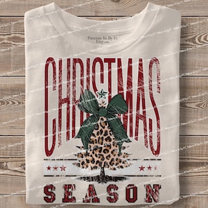 May include: Cream-colored t-shirt with a distressed red "CHRISTMAS SEASON" graphic. The design features a leopard print Christmas tree with a green bow and a star on top. Three stars are below the tree.