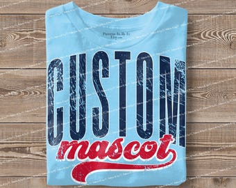 May include: Light blue t-shirt with the words "CUSTOM mascot" in distressed navy blue and red lettering. The shirt is folded on a wooden surface. The text has a vintage, worn look.
