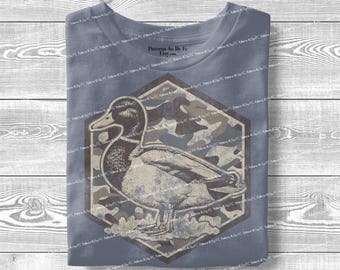 May include: A blue t-shirt featuring a detailed illustration of a duck within a hexagonal frame. The frame has a camouflage pattern. The duck is depicted in shades of brown and gray, with the shirt's background showing a faded blue hue.