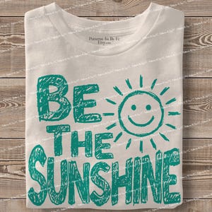 May include: A folded white t-shirt with the teal text "BE THE SUNSHINE" and a smiling sun graphic. The shirt is made of a soft material and is displayed on a wooden surface.