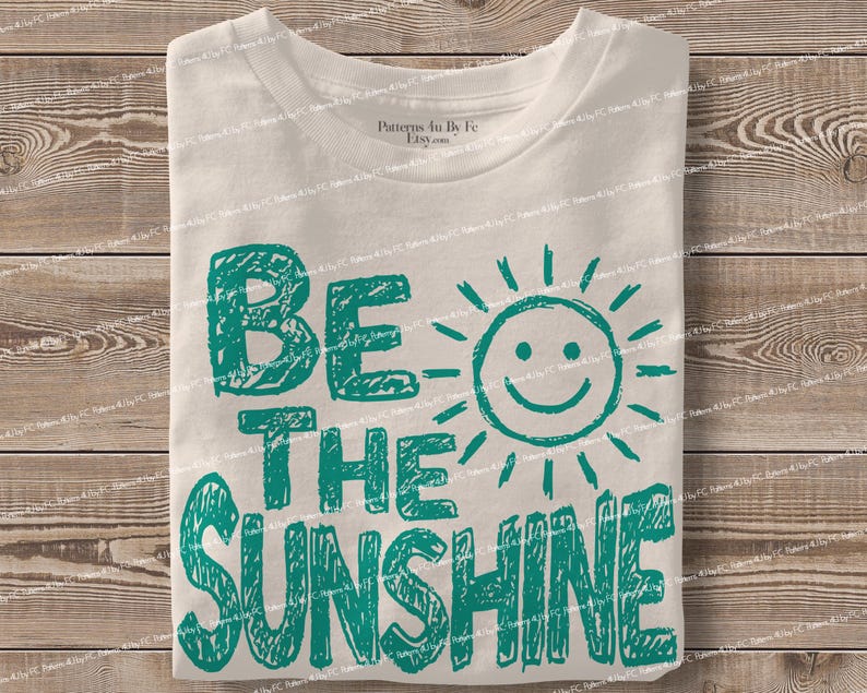 May include: A folded white t-shirt with the teal text "BE THE SUNSHINE" and a smiling sun graphic. The shirt is made of a soft material and is displayed on a wooden surface.