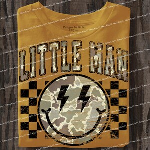 K&ouml;nnte beinhalten: Senfgelbes T-Shirt mit dem Text "LITTLE MAN" in einer verblassten Schriftart. Darunter befindet sich ein Smiley mit Blitzaugen und einem Tarnmuster. Der Hintergrund hat ein schwarz-gelbes Schachbrettmuster.