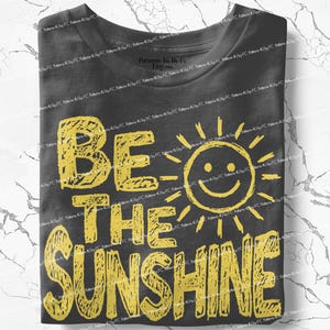 May include: A folded charcoal gray t-shirt with the words "BE THE SUNSHINE" in yellow block letters. A smiling sun graphic is next to the word "BE". The shirt is made of a soft material.