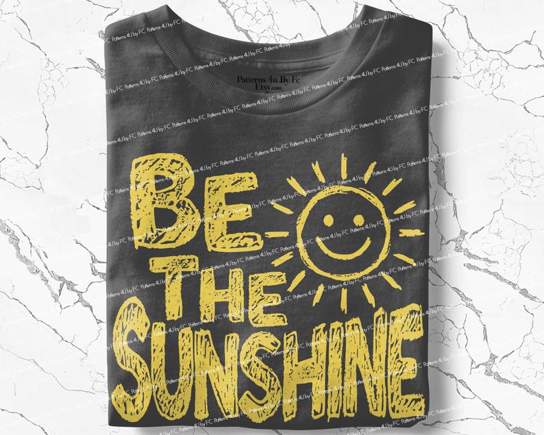 May include: A folded charcoal gray t-shirt with the words "BE THE SUNSHINE" in yellow block letters. A smiling sun graphic is next to the word "BE". The shirt is made of a soft material.