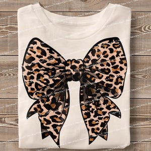 May include: A white t-shirt with a large brown and black leopard print bow on the front.