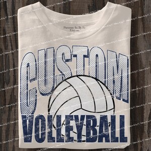 May include: Cream-colored t-shirt featuring the words "CUSTOM VOLLEYBALL" in a distressed navy blue font. A volleyball graphic is centered between the words. The shirt is folded and ready to wear.
