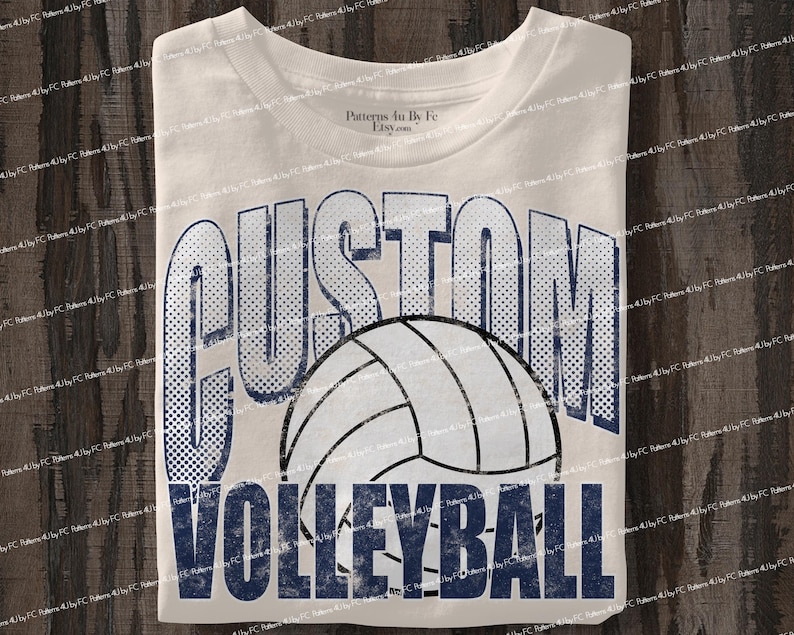 May include: Cream-colored t-shirt featuring the words "CUSTOM VOLLEYBALL" in a distressed navy blue font. A volleyball graphic is centered between the words. The shirt is folded and ready to wear.