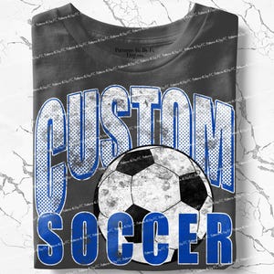 May include: Dark grey t-shirt featuring the words "CUSTOM SOCCER" in blue and white distressed lettering. A soccer ball graphic is centered below the word "CUSTOM". The shirt is folded.