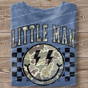 K&ouml;nnte beinhalten: Blaues T-Shirt mit dem Text "LITTLE MAN" in einer verblassten Schriftart. Darunter ein Smiley mit Camouflage-Muster und Blitzaugen, vor einem schwarz-wei&szlig; karierten Hintergrund.