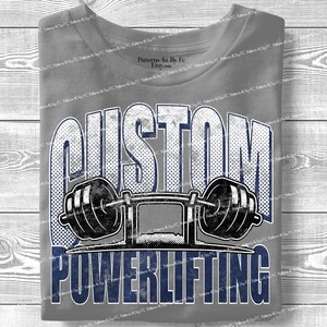 Könnte beinhalten: Ein graues T-Shirt mit dem Text "CUSTOM POWERLIFTING" in einem Used-Design in Marineblau und Weiß. Eine Hantel-Grafik ist unter dem Wort "CUSTOM" zentriert. Das Shirt ist sauber auf einer Holzoberfläche gefaltet.