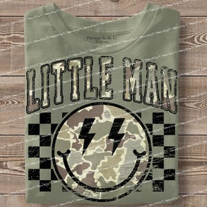 K&ouml;nnte beinhalten: Olivgr&uuml;nes T-Shirt mit dem Schriftzug "LITTLE MAN" in einer verwaschenen Schriftart. Darunter befindet sich ein Smiley mit Blitzaugen und einem Tarnmuster. Der Hintergrund hat ein schwarz-wei&szlig;es Schachbrettmuster.
