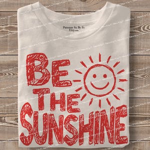 May include: A folded white t-shirt with the red text "BE THE SUNSHINE" and a smiling sun graphic. The shirt is made of a soft material and is folded neatly on a wooden surface.